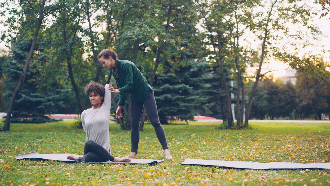 Two adult women practicing yoga in a park, with an instructor assisting one of them with a pose.