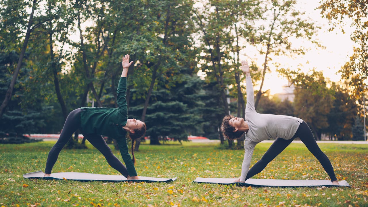 Two women practicing yoga outdoors on a grassy park at sunset.