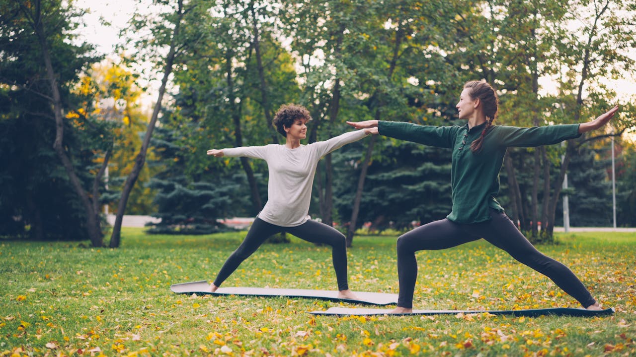 Two women practicing yoga outdoors on mats in a park during fall.